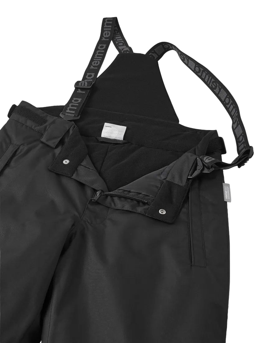 Close-up view of black Reima Jr Terrie Pants featuring a waterproof Reimatec shell, detachable elastic suspenders, reinforced ankle scuff guards, and internal snow gaiters.