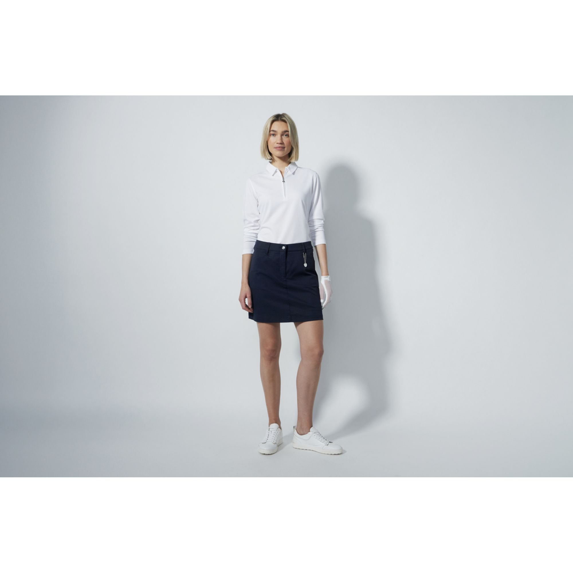 A woman wearing a white Daily Sports shirt and navy Daily Sports golf skort standing against a grey background.
