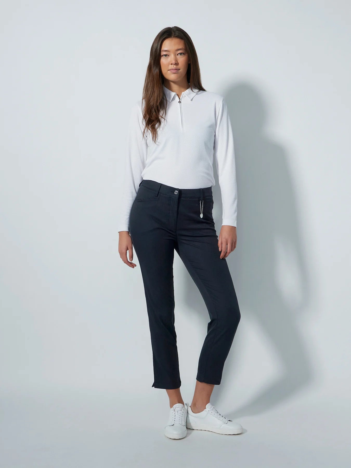 A woman modeling a pair of dark gray, high-waisted, ankle-length daily sports golf pants with a small slit at the ankle, an elastic waistband, and stretchy fabric. She is also wearing a white long-sleeved shirt and white sneakers.