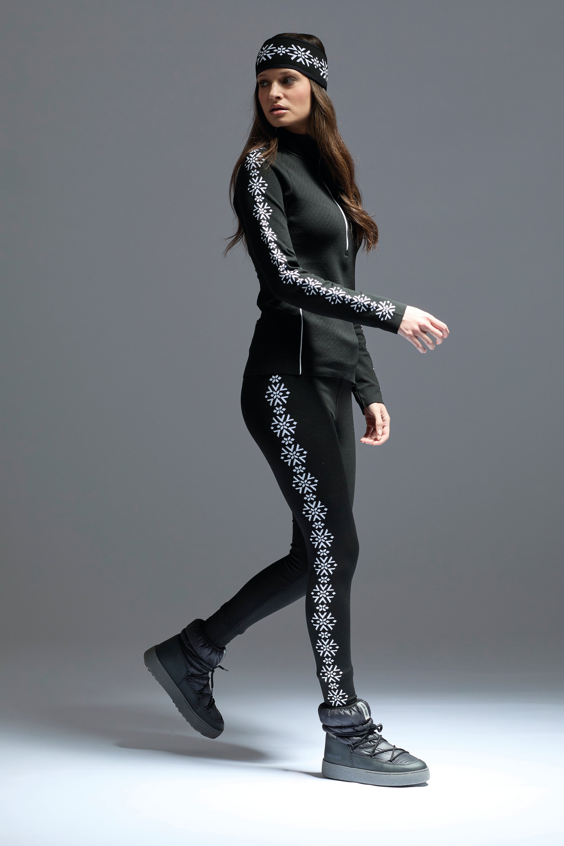 A woman wearing a pair of black Newland Women's Croix de Couer Leggings that feature a white snowflake pattern down the legs and are moisture-wicking, breathable, and stretchable.