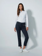 A woman modeling a pair of dark gray, high-waisted, ankle-length daily sports golf pants with a small slit at the ankle, an elastic waistband, and stretchy fabric. She is also wearing a white long-sleeved shirt and white sneakers.