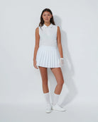 Woman wearing a Daily Sports white sleeveless top and pleated skirt with white shoes on a plain background