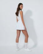 Woman wearing a Daily Sports white sleeveless top and pleated skirt with white shoes on a plain background