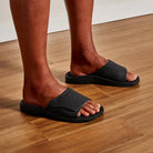 A man wearing a pair of black Olukai Men's Nalu Slide sandals with a strap on hardwood flooring