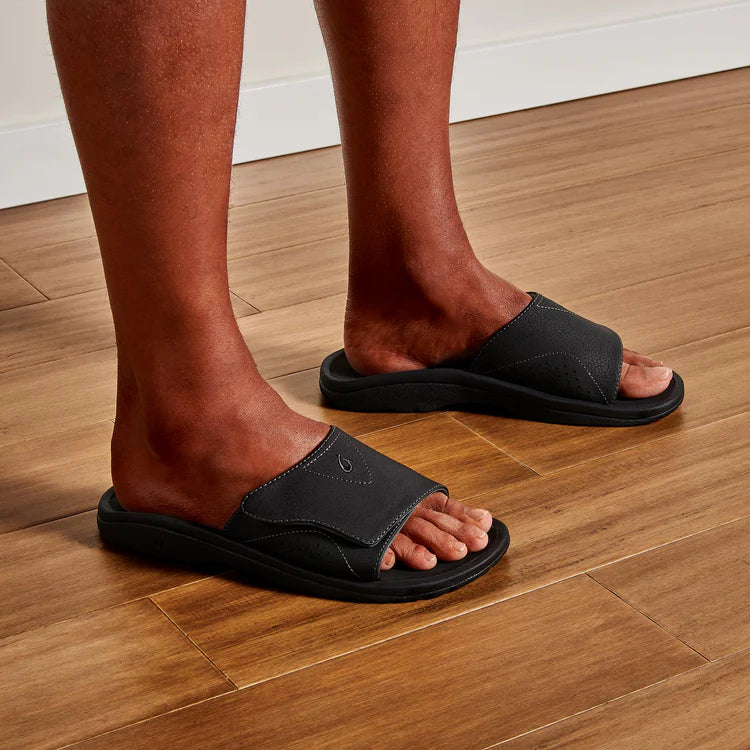 A man wearing a pair of black Olukai Men's Nalu Slide sandals with a strap on hardwood flooring