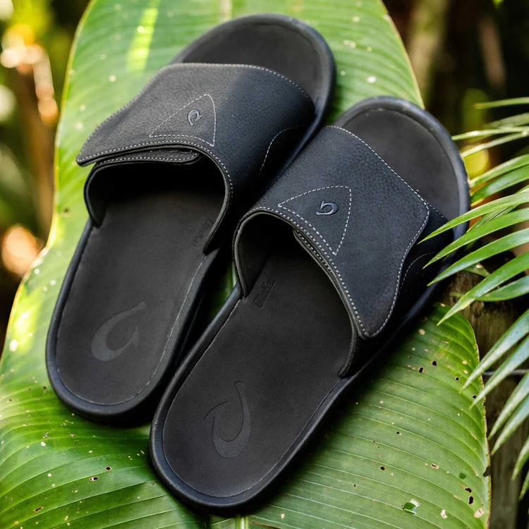 A pair of black Olukai Men's Nalu Slide sandals with a strap on a tropical leaf