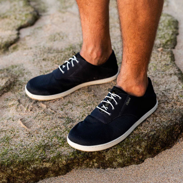 A man wearing a pair of black Olukai Men's Nohea Moku shoes standing on a rock on the beach