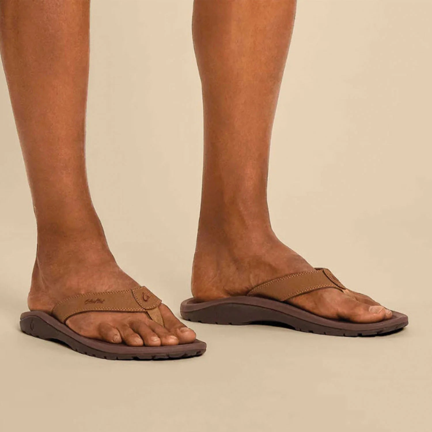 A man wearing a pair of brown Olukai Mens Nui Sandals on a beige background