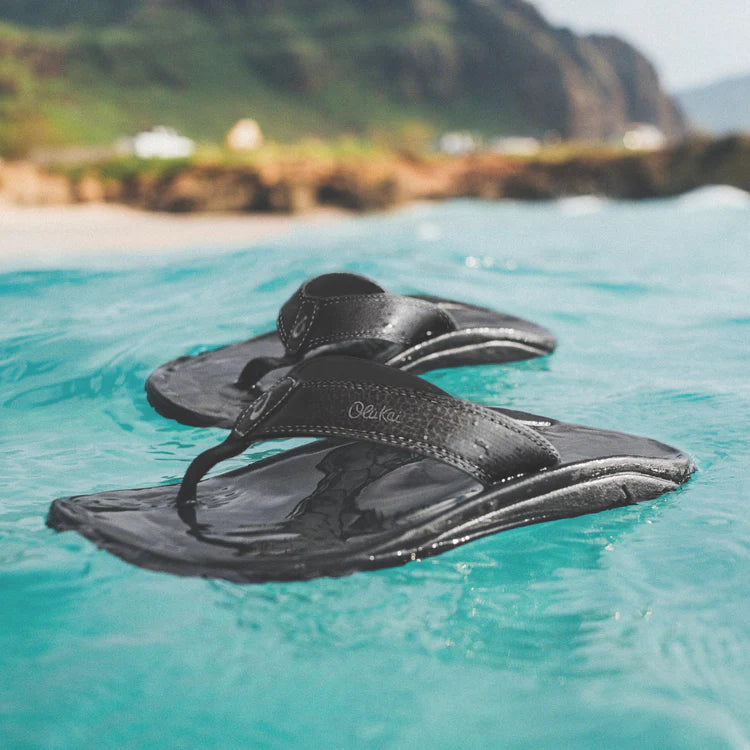 A pair of black Olukai Men's Ohana flip-flop sandals with an Olukai logo floating on water