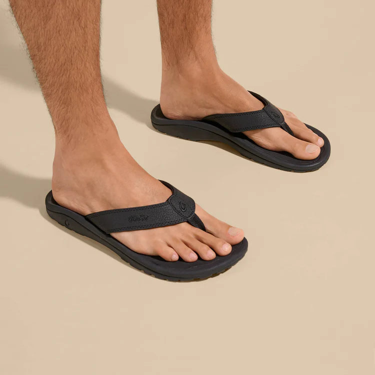 A man wearing a pair of black Olukai Men's Ohana flip-flop sandals with an Olukai logo on a beige background