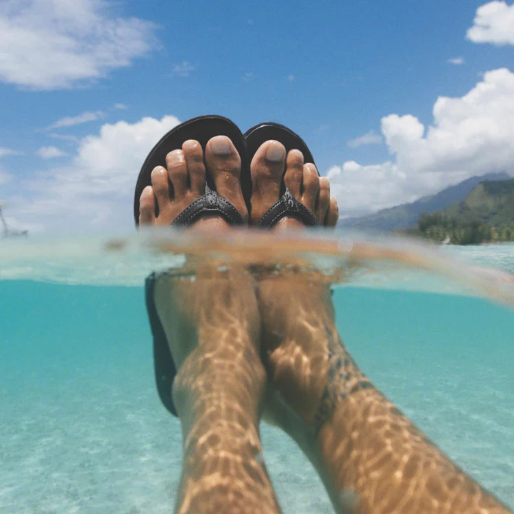 A woman wearing black Olukai Women's Ho'opio flip-flop sandals with feet submerged in clear blue water, surrounded by a tropical landscape.