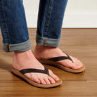 A woman wearing a pair of brown leather Olukai Women's Nonohe flip-flop sandals