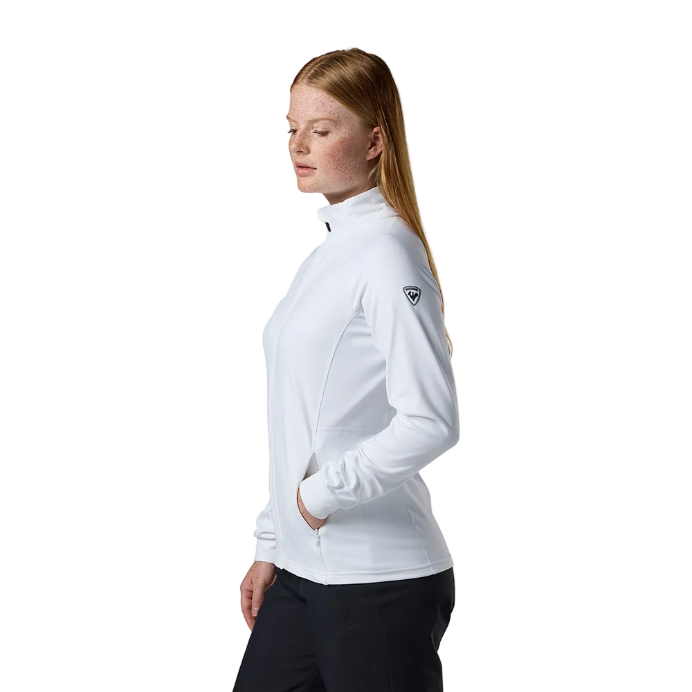 Side view of a white Rossignol Women’s Midlayer Stretch Jacket featuring multi-directional stretch fabric, a thermal fleece interior, full-length front zipper, and zippered hand pockets.