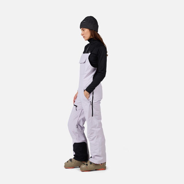 A woman wearing a pair of light purple Rossignol Women's Outer Limits Insulated Bib Ski Pants that are waterproof, breathable, stretchable, and warm.