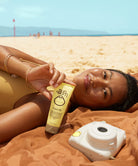 Woman on a beach with a Sun Bum Original SPF 50 Sunscreen Face Lotion bottle and camera nearby
