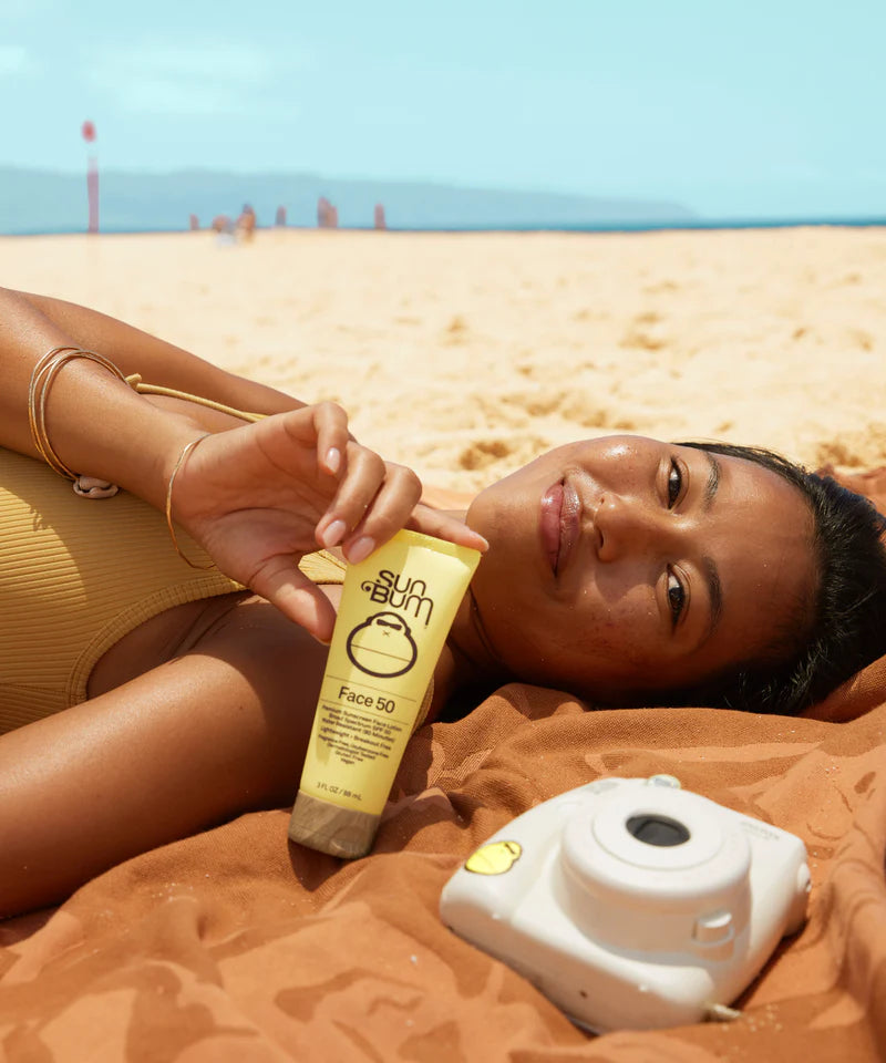Woman on a beach with a Sun Bum Original SPF 50 Sunscreen Face Lotion bottle and camera nearby